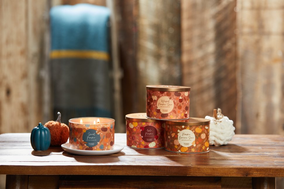 the pumpkin patch collection featuring spiced pumpkin 3 wick candle, whipped pumpkin spice 3 wick candle, apple pumpkin 3 wick candle, and pumpkin banana scone 3 wick candle on a wood table with white, blue, and orange decorative pumpkins