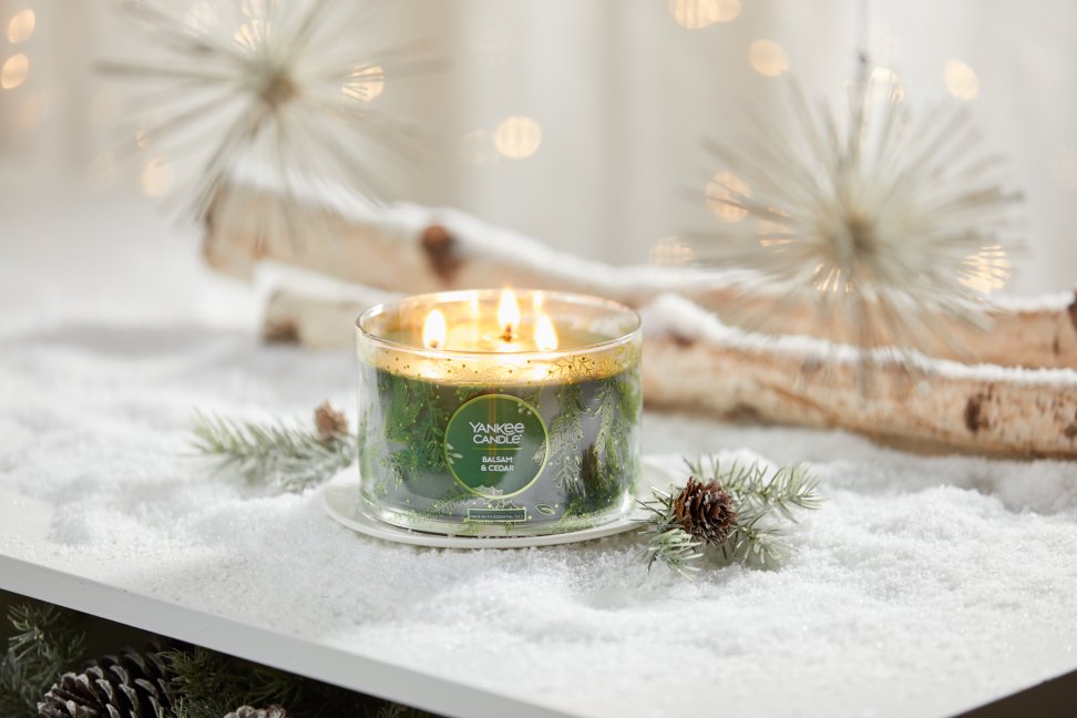 balsam and cedar three wick candle on plate on snowy surface with pinecones evergreens and birch branches