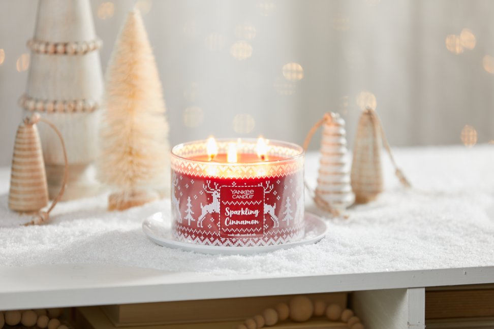 sparkling cinnamon three wick candle on white plate on snowy surface with rustic wood and brush christmas trees