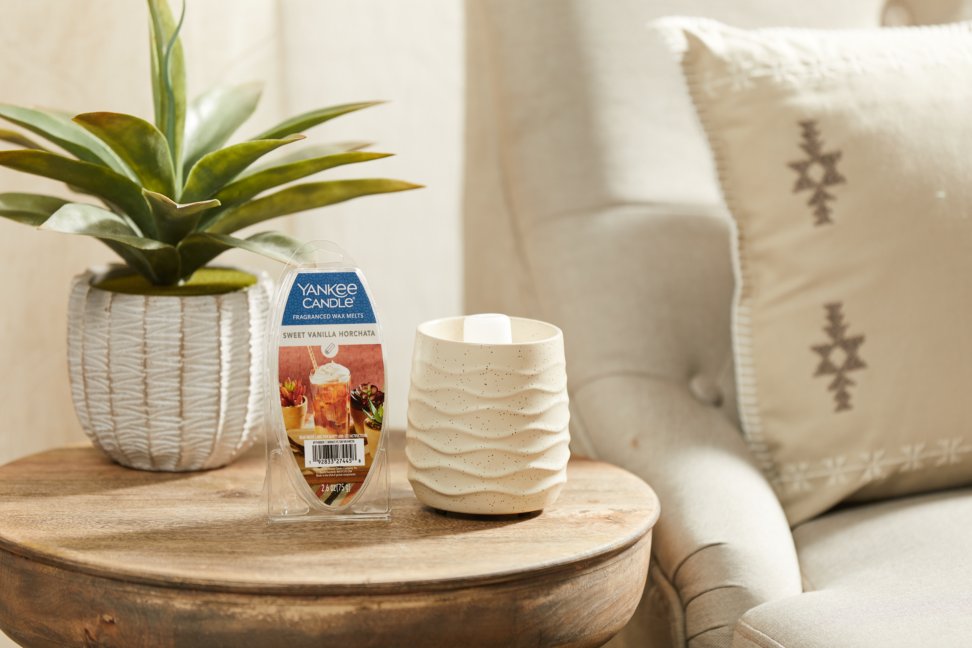 a plant in a white pot next to a sandstone electric wax melt warmer and sweet vanilla horchata wax melts on a wood side table next to a white couch with a decorative pillow