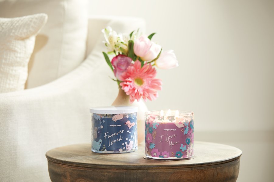 an "I Love You" 3-Wick candle and a "Furever loved" 3-Wick candle on a table next to a vase of flowers