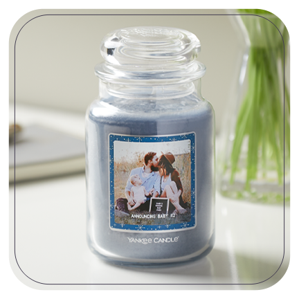 a blue-colored original large jar candle with a personalized photo label with a man, a woman, and a young child and announcing baby #2 messaging on a white table
