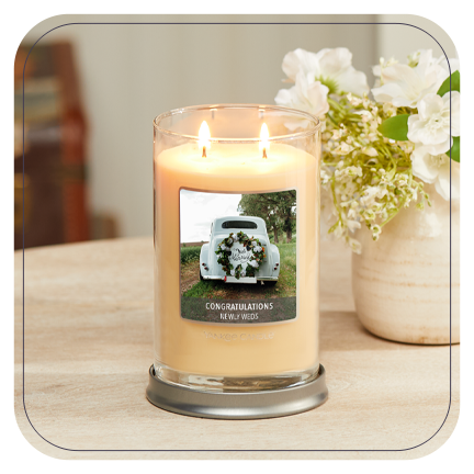 a cream-colored signature large tumbler candle with a personalized photo label of a white car and messaging reading congratulations newly weds on a wood table next to a vase filled with white flowers